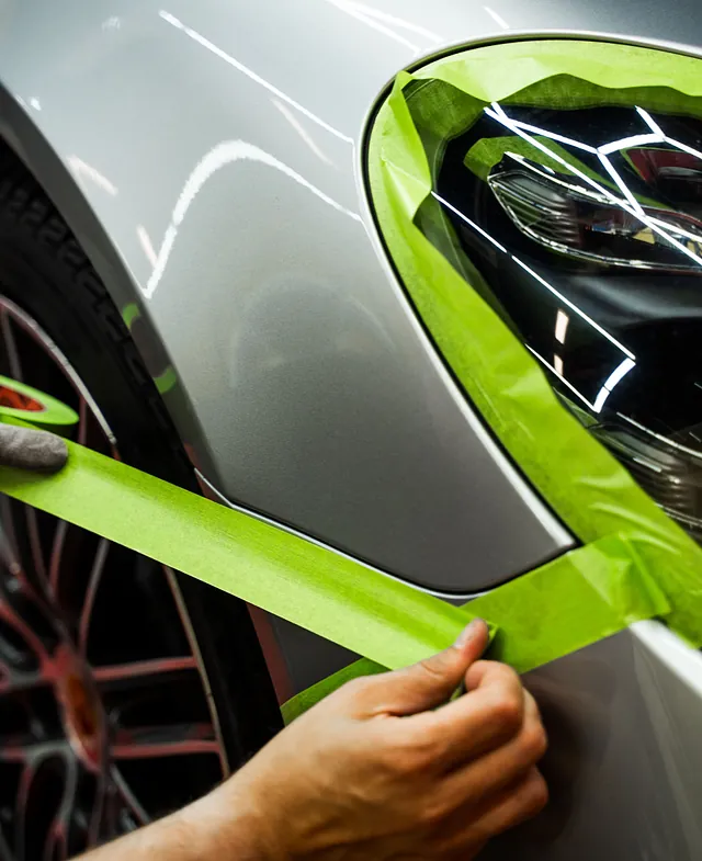 Person som legger grønn maskeringstape rundt frontlykten på en grå bil før arbeid på lakken.