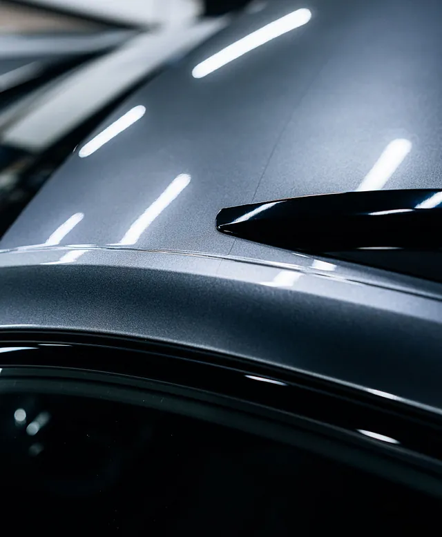 Detaljbilde av taket på en grå bil med en svart takspoiler og refleksjoner fra lys i lakken.
